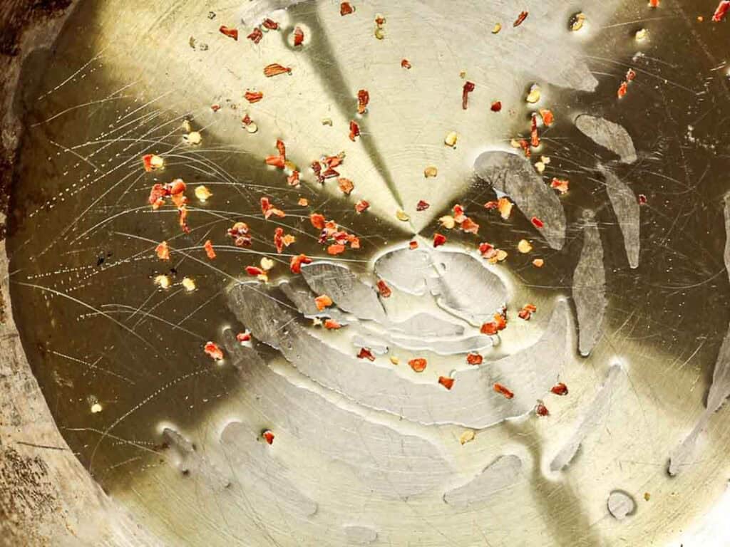 A close-up of a scratched metal surface with scattered red chili flakes and oil droplets, creating a textured and reflective appearance.